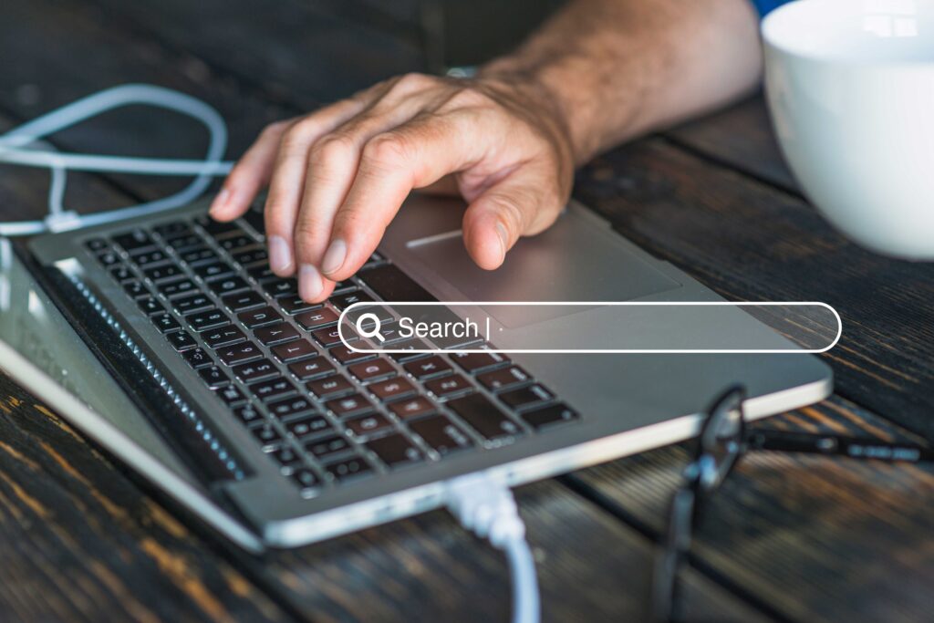 Close-up of a hand typing on a laptop keyboard, with white cables and a mug on a dark wooden desk; a 'Search' bar overlay on screen.